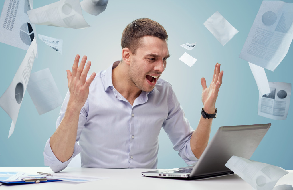 business, people, stress, fail and technology concept - angry businessman with laptop computer shouting over blue background and falling papers